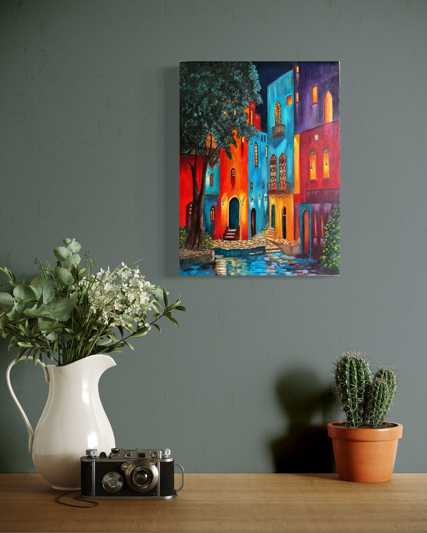 Colorful painting of a street scene on a wall above a table with a pitcher, camera, and plants.